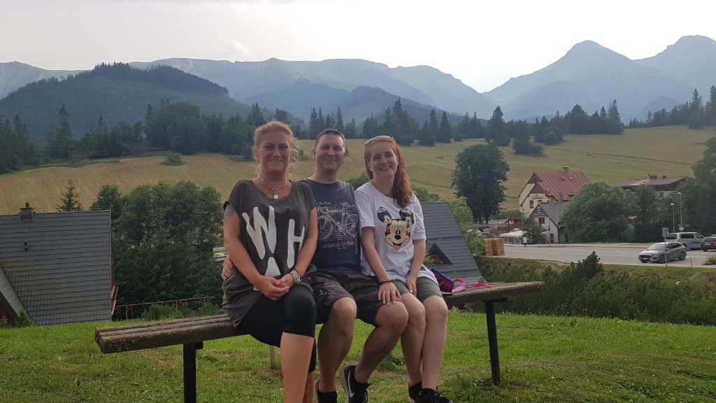 Traja ľudia na drevenej lavičke s panorámou hôr v pozadí.