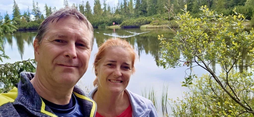 Selfie páru pred tichým jazerom a ihličnatým lesom.